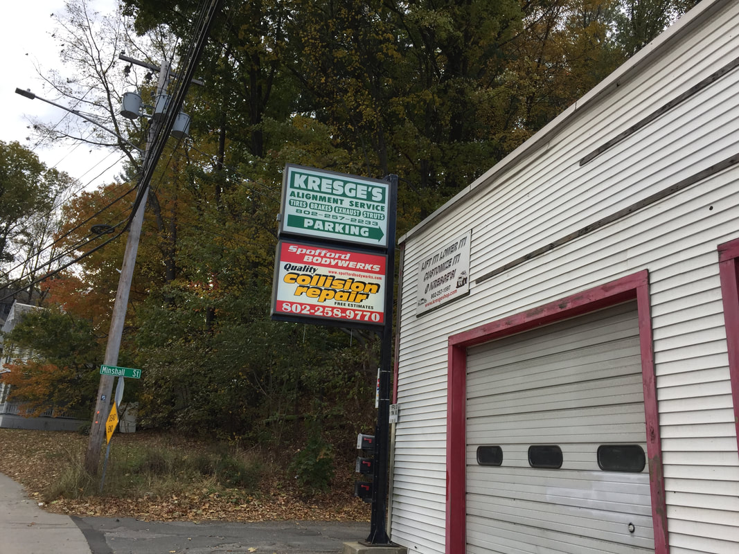 Spofford Bodywerks shop on Canal Street — red garage door and the roadside ‘Quality collision repair · Free estimates’ sign, autumn foliage behind